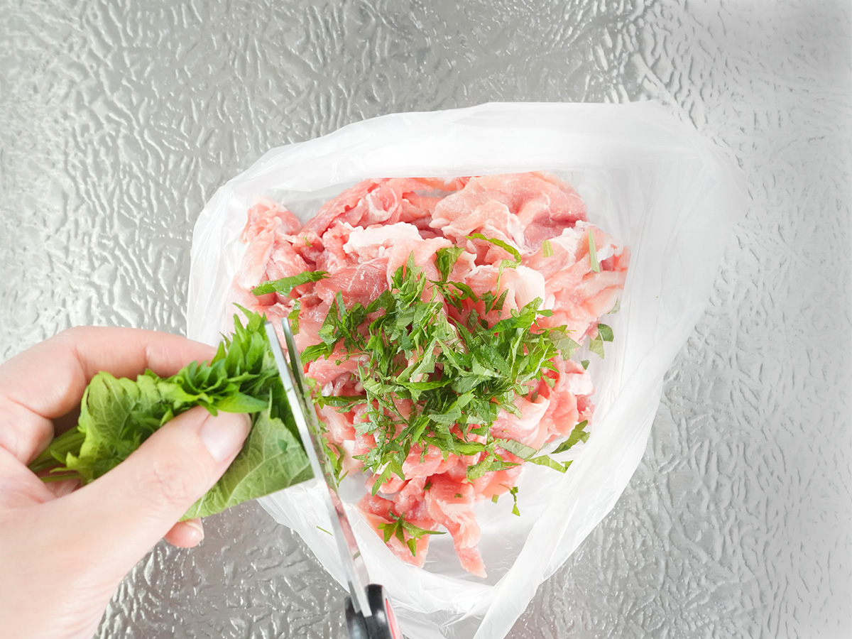 ポリ袋に豚こま肉を入れ、大葉をはさみで切って入れるシーン
