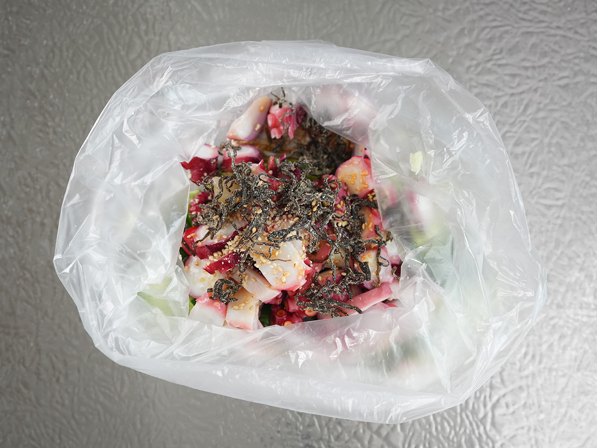 きゅうりが入ったポリ袋に茹でダコを入れて、Aのごま油、鶏がらスープの素、しょうゆ、にんにく、塩昆布、白ごまを加えるシーン